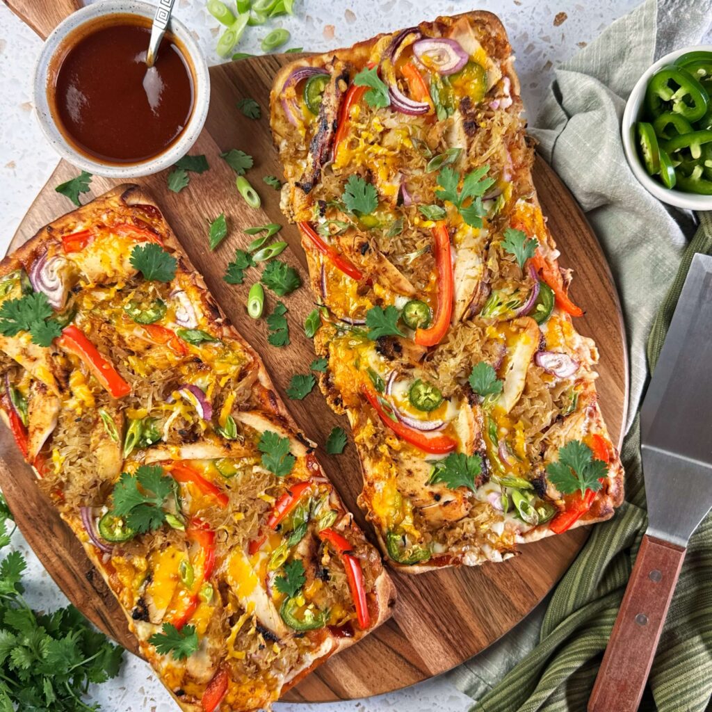 Two BBQ Kraut and Chicken Flatbreads on a round wooden board with a cup of sauce.