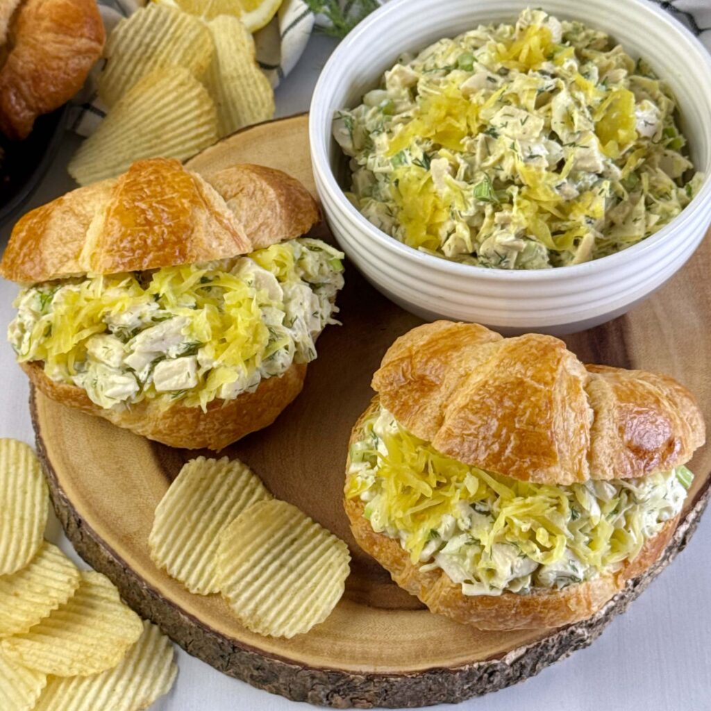 Two Mustard Kraut Chicken Salad Sandwiches on a round wooden board with a white bowl with extra Mustard Kraut and Chicken Salad