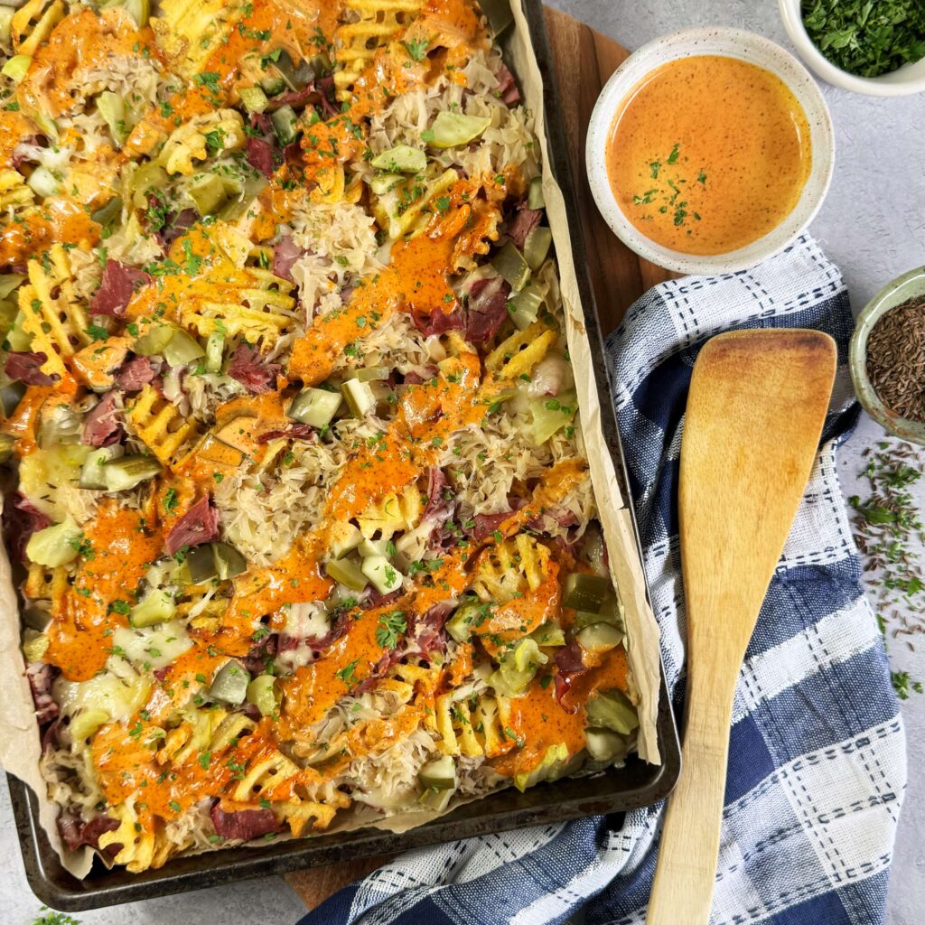 A baking sheet with Loaded Reuben Waffle Fries with Zesty Thousand Island Dressing on top on a counter display with a wooden spoon and a side bowl of Zesty Thousand Island dressing.