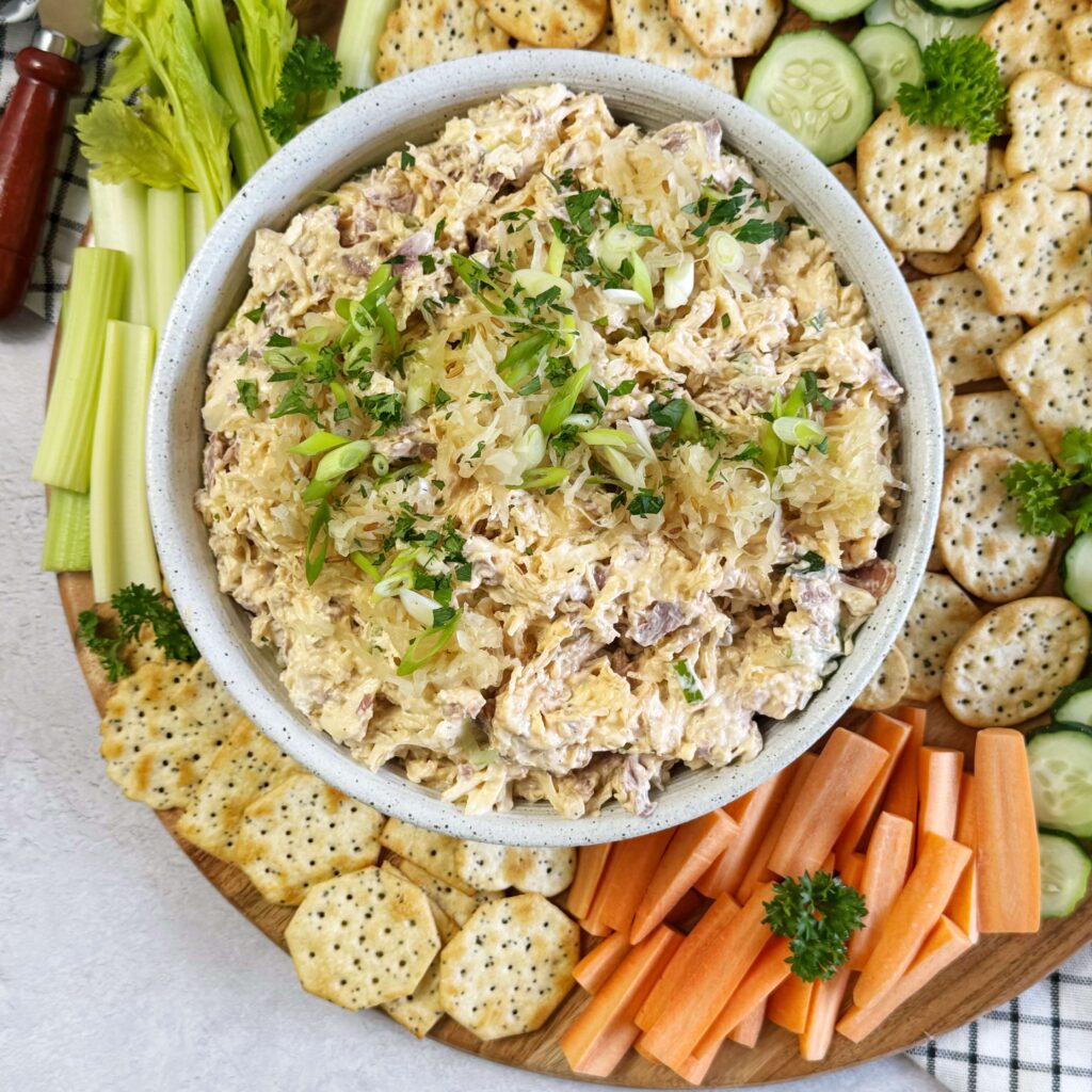 A white bowl of Reuben Cracker Spread with Bavarian. On a wooden tray with crackers, carrots, celery and cucumbers.
