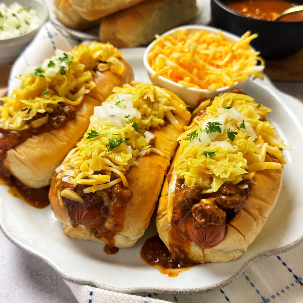 A white plate with three Chile Cheese Hot Dogs on the plate loaded with Mustard Kraut, onion and cheese.