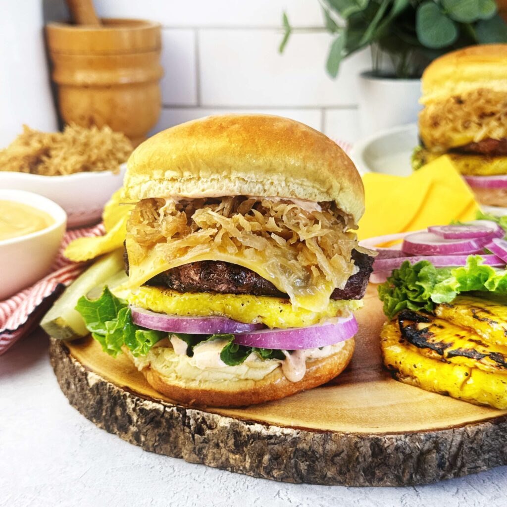 Juicy grilled burger topped with grilled pineapple, BBQ kraut, and BBQ mayonnaise on a toasted bun, served as a flavorful summer cookout meal.